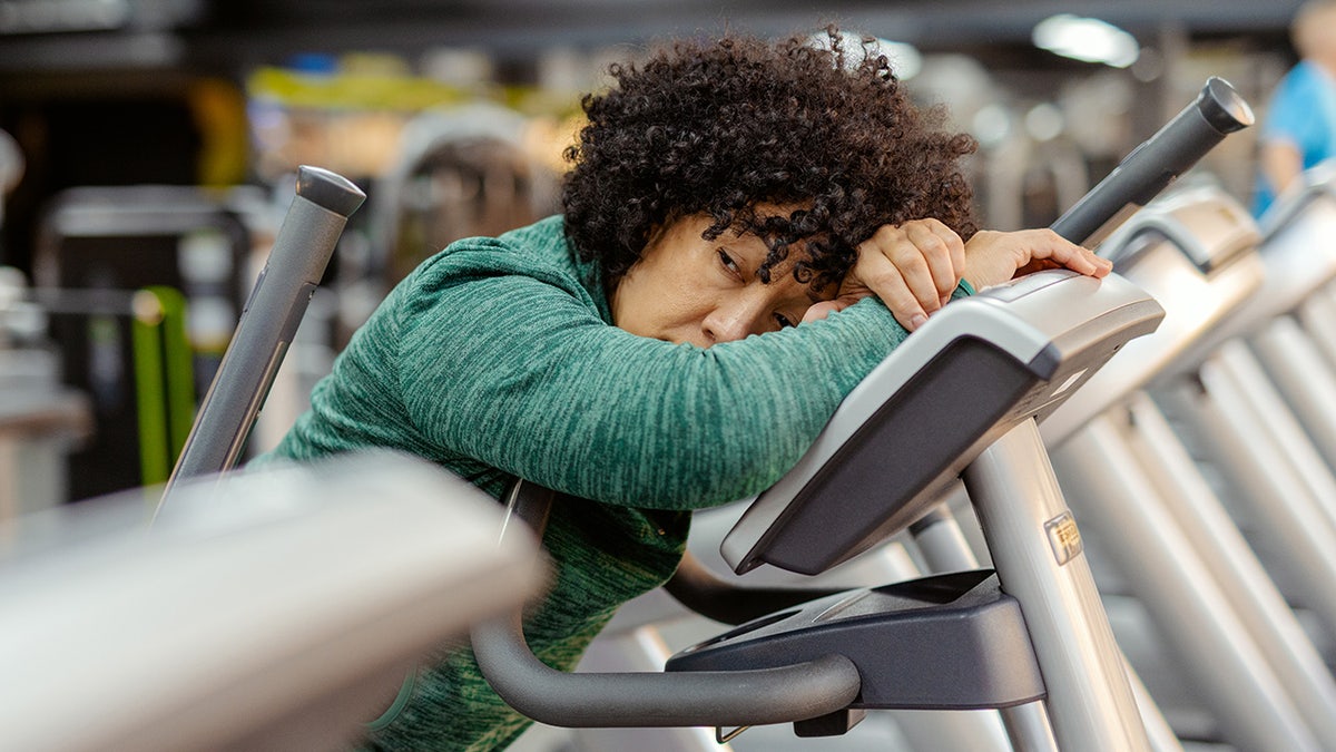 Middle aged woman taking a break, head down on treadmill at gym, looking exhausted.
