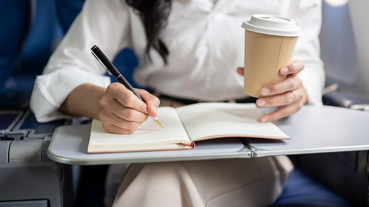 Woman writing in notebook with a pen while drinking coffee on a plane.