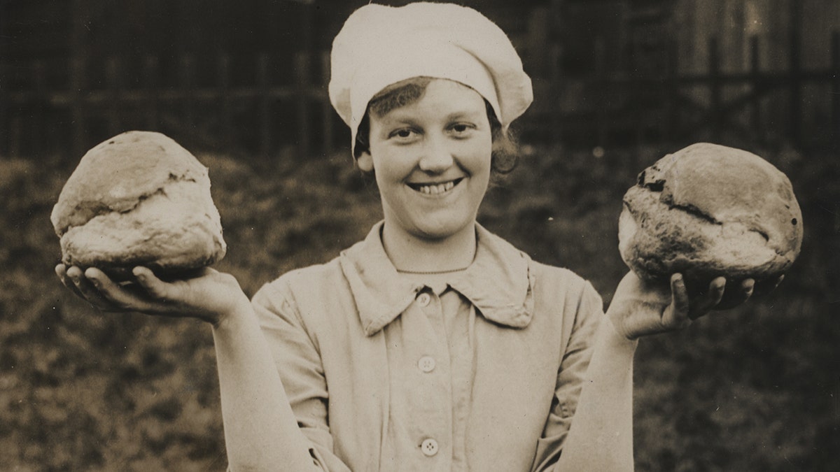 Woman holding freshly baked bread during WWI, seen outside, smiling and posing with loaf in each hand and wearing a chef hat.