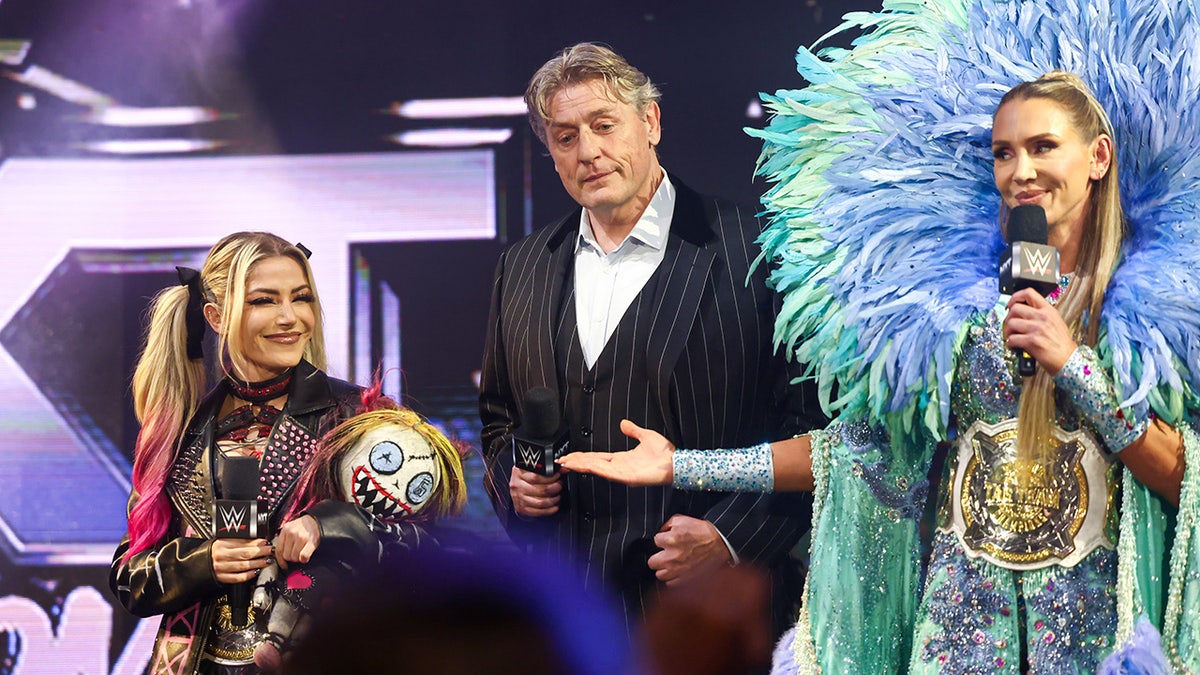 William Regal in between Alexa Bliss and Charlotte Flair