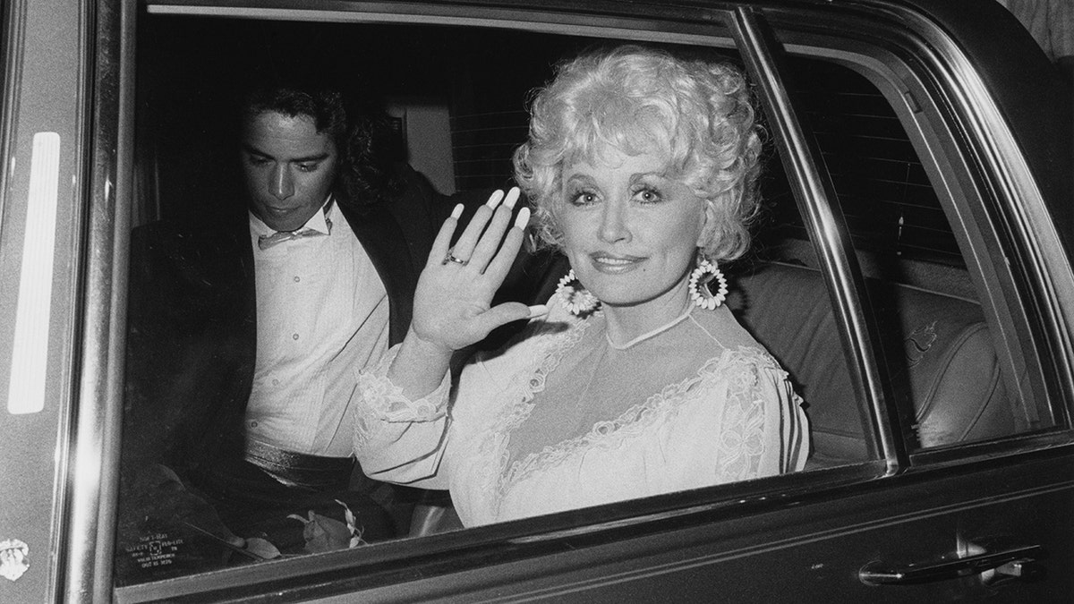 A woman is waving her hand as she sits in the backseat of a car with a man sitting next to her.