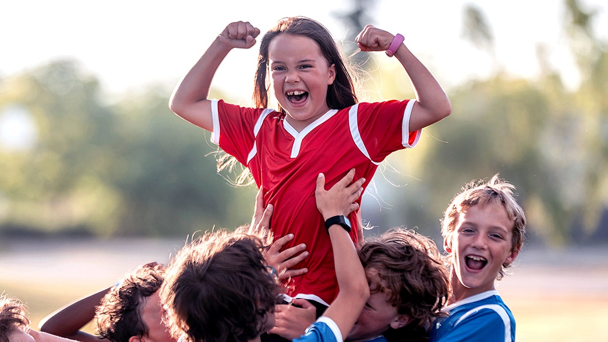 A joyful youth soccer team celebrates their victory as teammates lift a smiling player skyward outdoors.