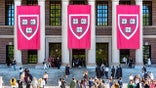 Three large “Veritas” banners hang from the front of Harvard’s Widener Memorial Library as graduating students pose for photos in their academic regalia.