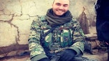 Man in camouflage uniform sits against a cracked wall while holding a rifle.