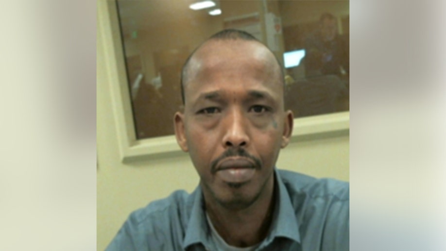 A man sits indoors facing the camera in a room with fluorescent lighting and a windowed partition behind him.