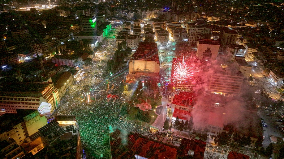 Crowds gather in a public square at night as fireworks and spotlights light up the sky for a political anniversary event.