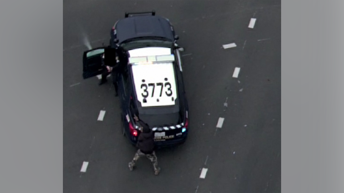 An aerial view shows a person standing behind a police vehicle with its door open