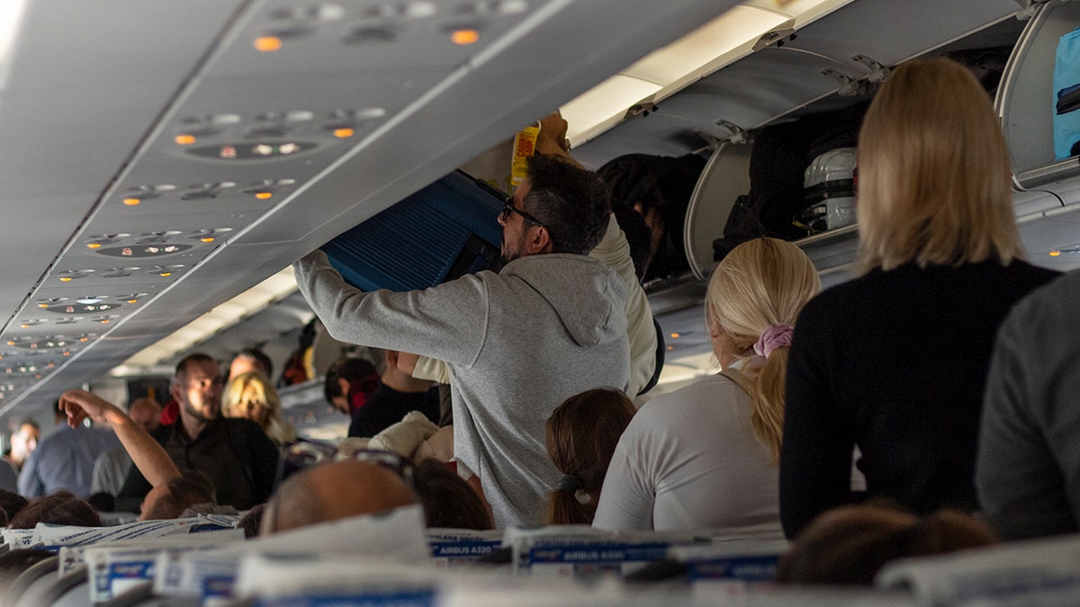 Passengers standing in a crowded airplane aisle while loading carry-on luggage into overhead bins.