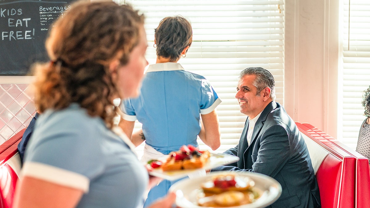 Waitress walks with breakfast plates in hand at diner while another waitress, seen from behind, talks with customer at table.