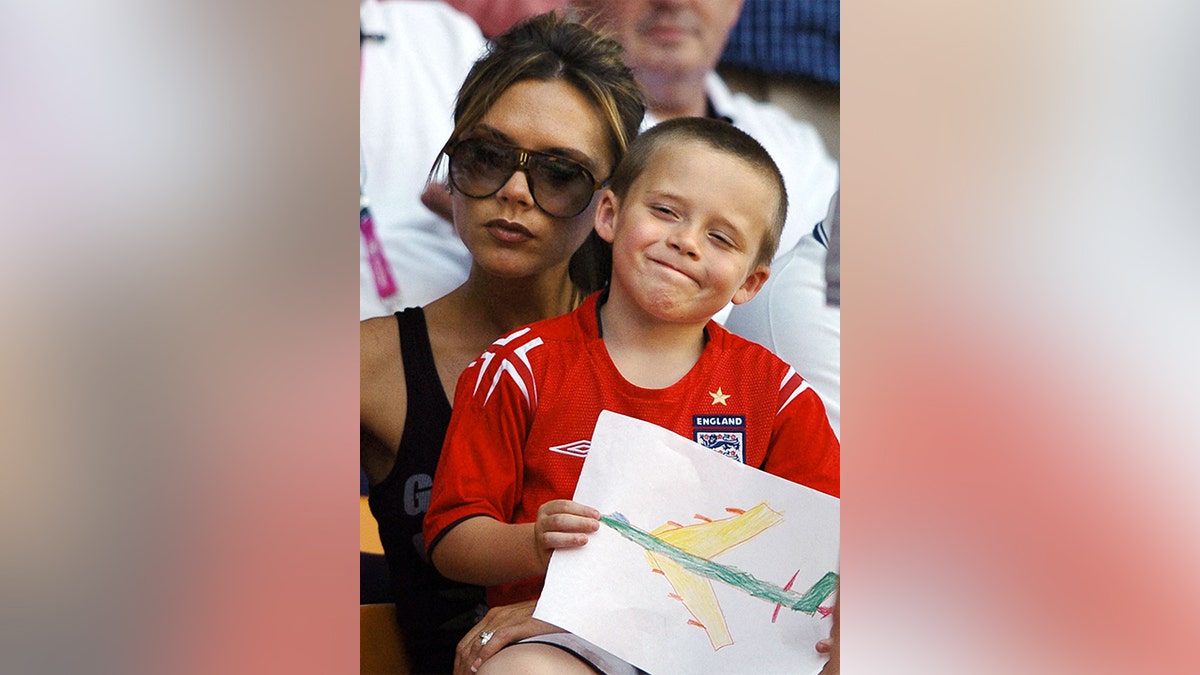 Brooklyn Beckham sitting on his mom Victoria Beckham's lap as they watch David Beckham play soccer.