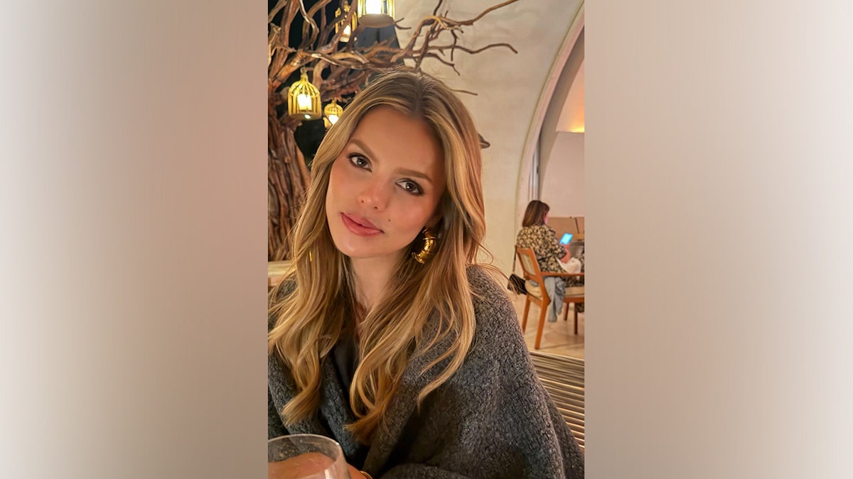 Brooks Nader sits indoors at a restaurant wearing a gray wrap and gold hoop earrings while dining out during her Cabo San Lucas vacation.
