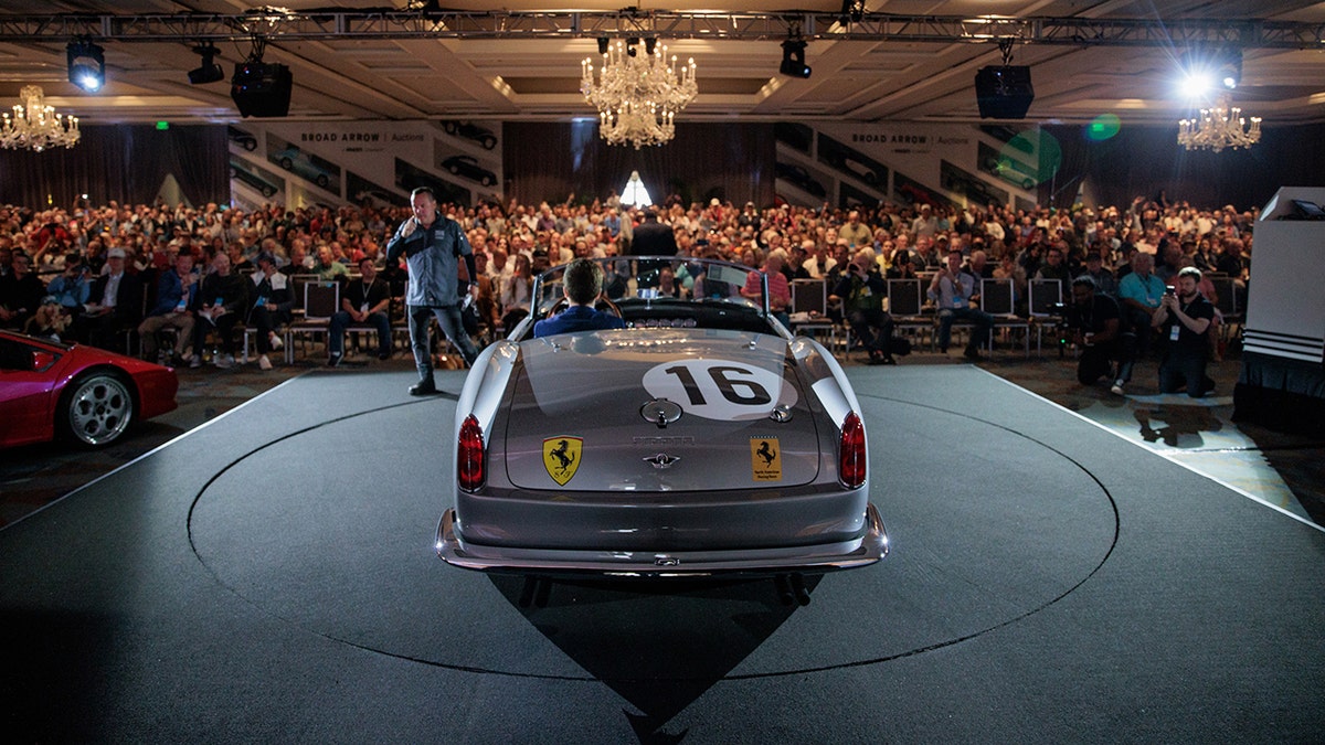 Vintage Ferrari race car displayed on stage at a live automotive auction with a large audience in attendance.