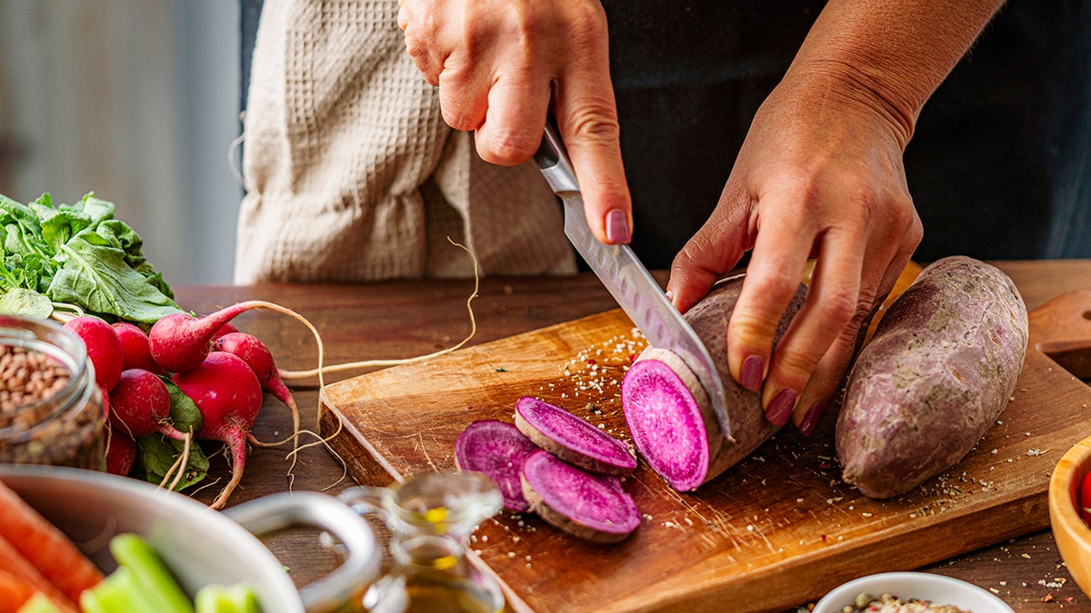 A woman cuts purple potatoes on a cutting board, and other produce such as tomatoes and radishes sit on the counter.