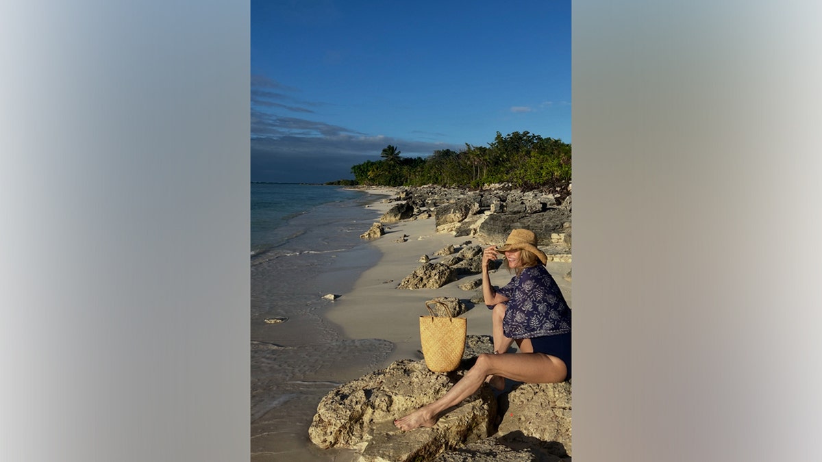 Christie Brinkley sits on a rocky shoreline during a tropical vacation, wearing a navy one-piece swimsuit, patterned cover-up and straw sun hat beside the ocean.