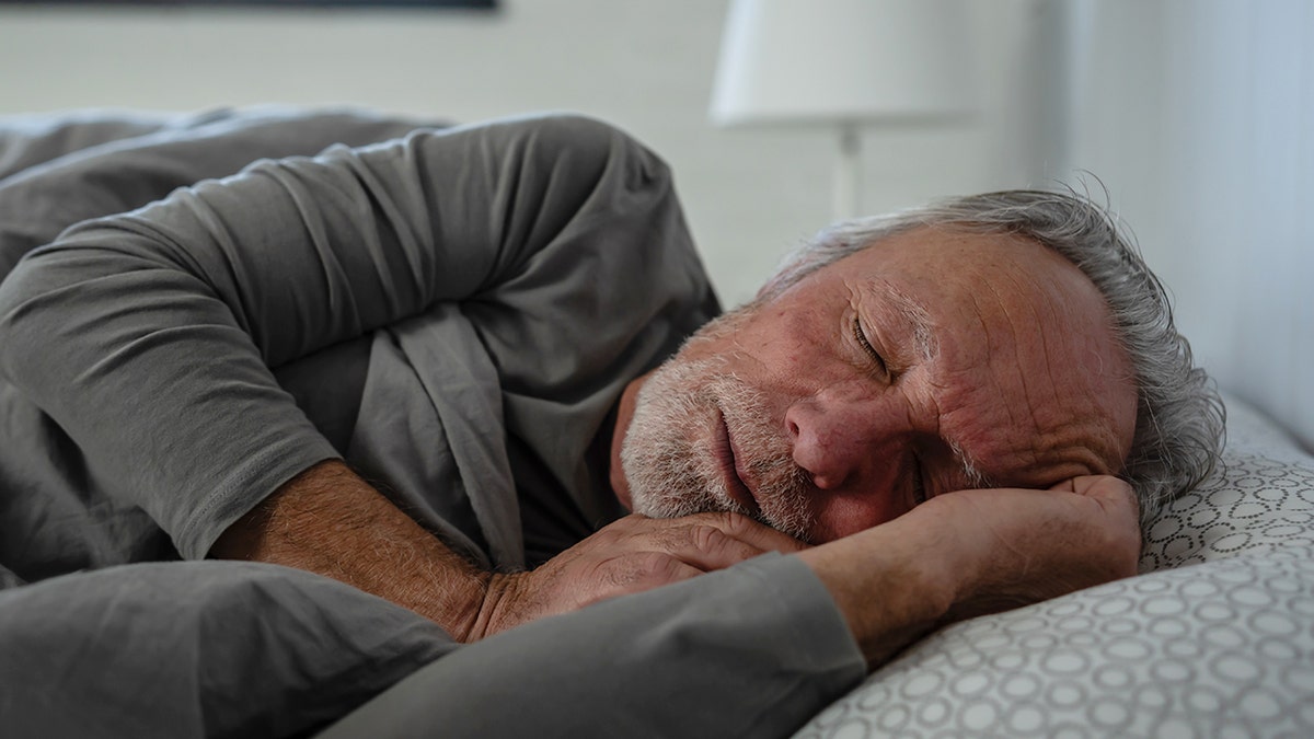 Man sleeping peacefully in bed, resting on his side under a gray blanket in a softly lit bedroom