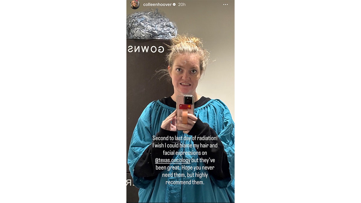 Author Colleen Hoover takes a mirror selfie in a medical setting, wearing a loose blue medical gown with her hair in a messy bun, holding a phone, with on-screen text mentioning it is her second-to-last day of radiation treatment.
