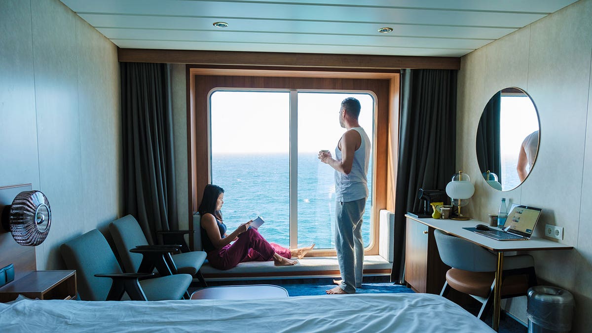Casal relaxando dentro de uma cabine de navio de cruzeiro com grande janela com vista para o mar.