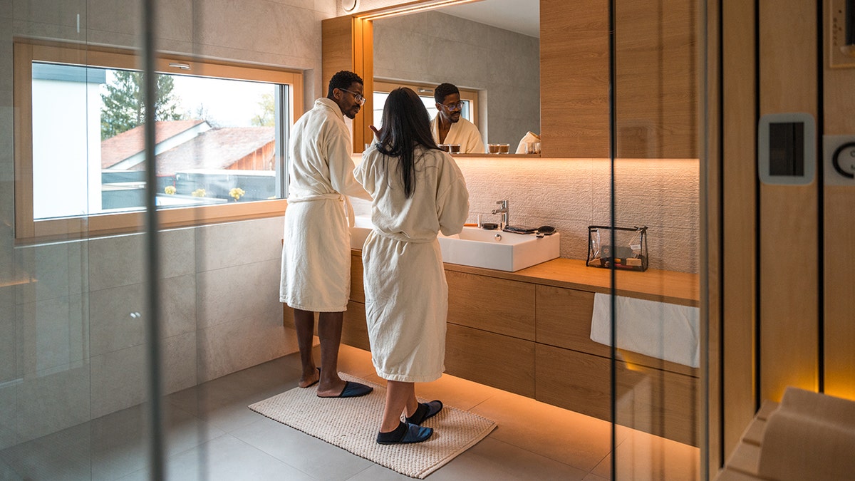 Couple in bathrobes standing at a modern bathroom vanity with a large mirror and warm wood finishes.