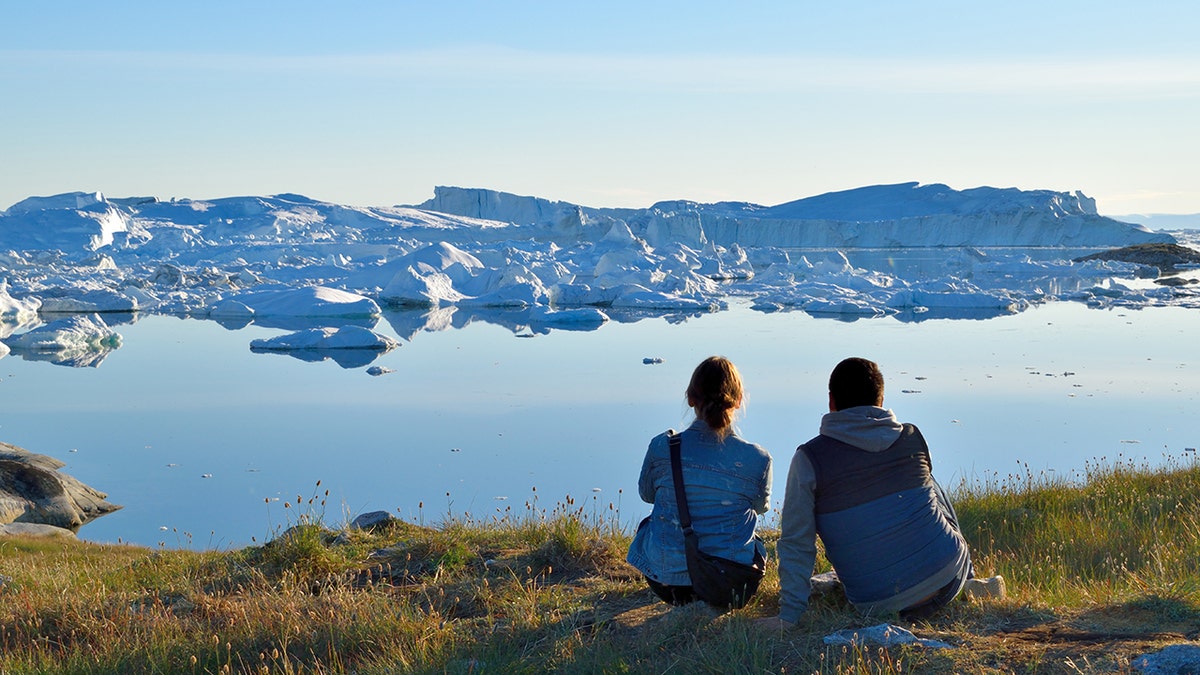 Two people sit on a grassy overlook facing a calm Arctic fjord filled with icebergs under a clear blue sky.