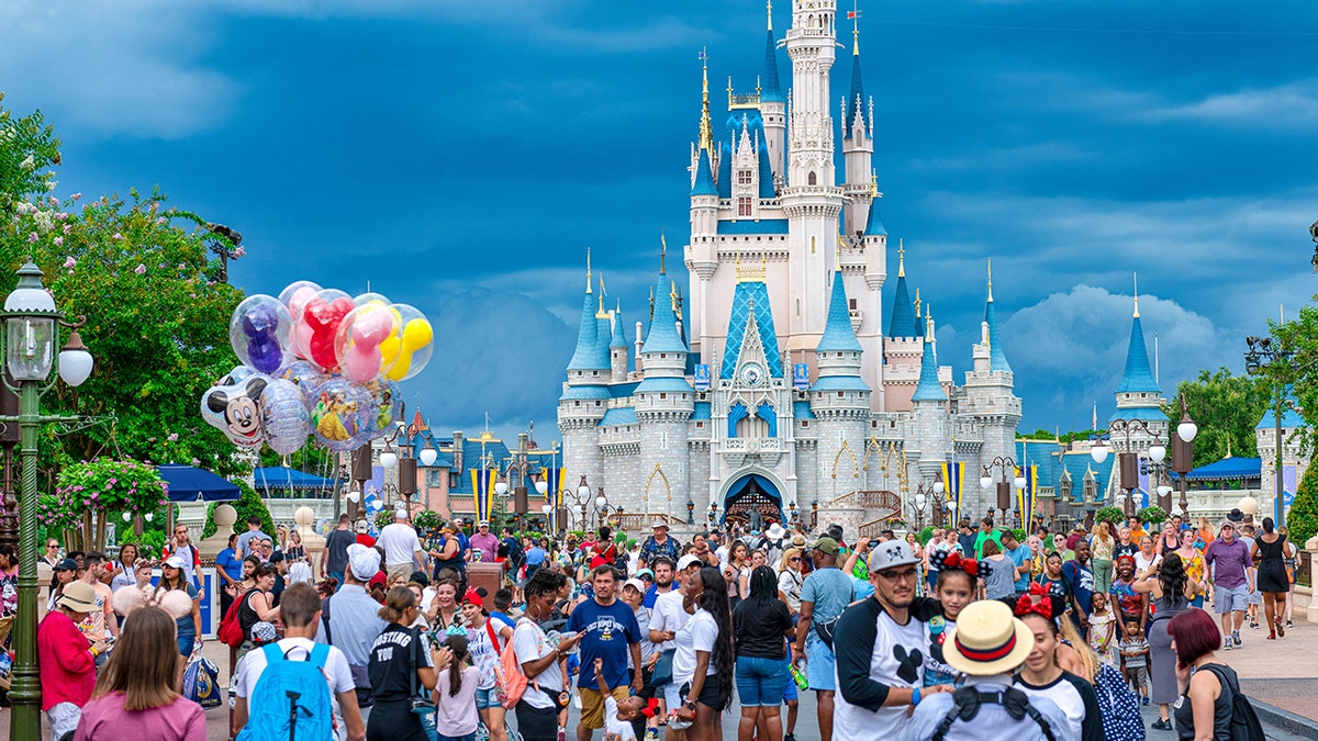 Crowds walking down street toward Cinderella Castle at Walt Disney World with colorful Mickey balloons under blue sky.