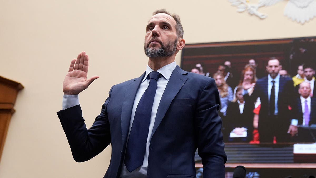 Jack Smith standing with his right hand raised while taking an oath in a committee room.