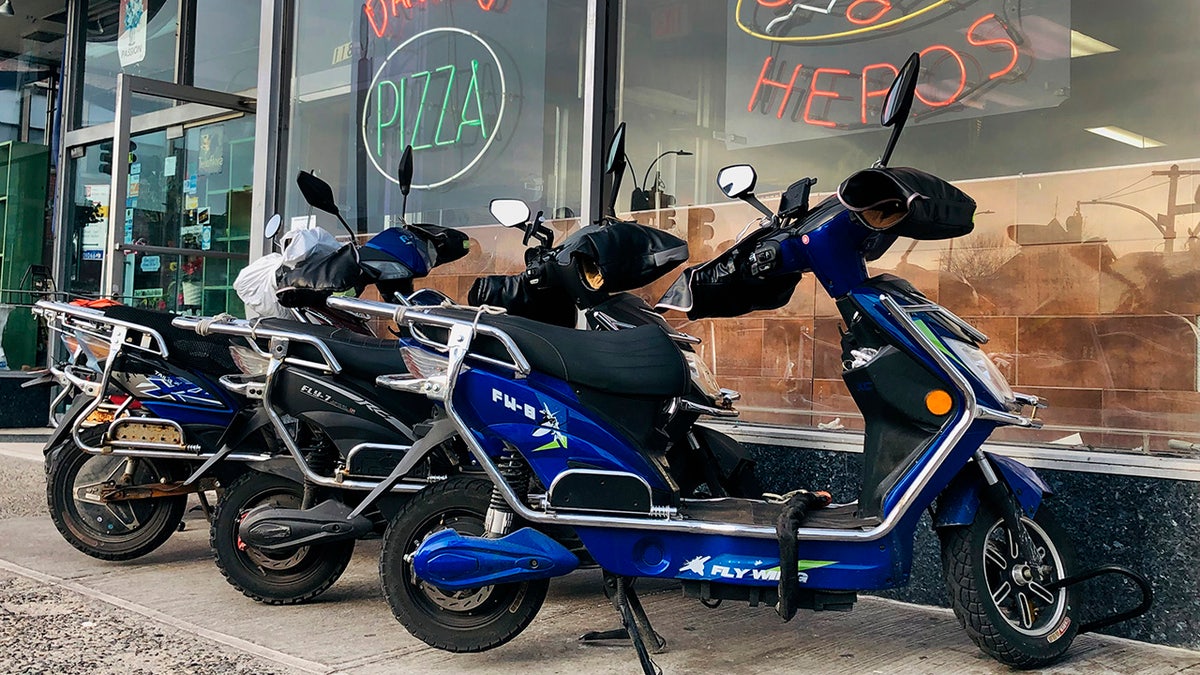 Electric scooters for delivery lined up on sidewalk outside NYC pizza and hero restaurant.
