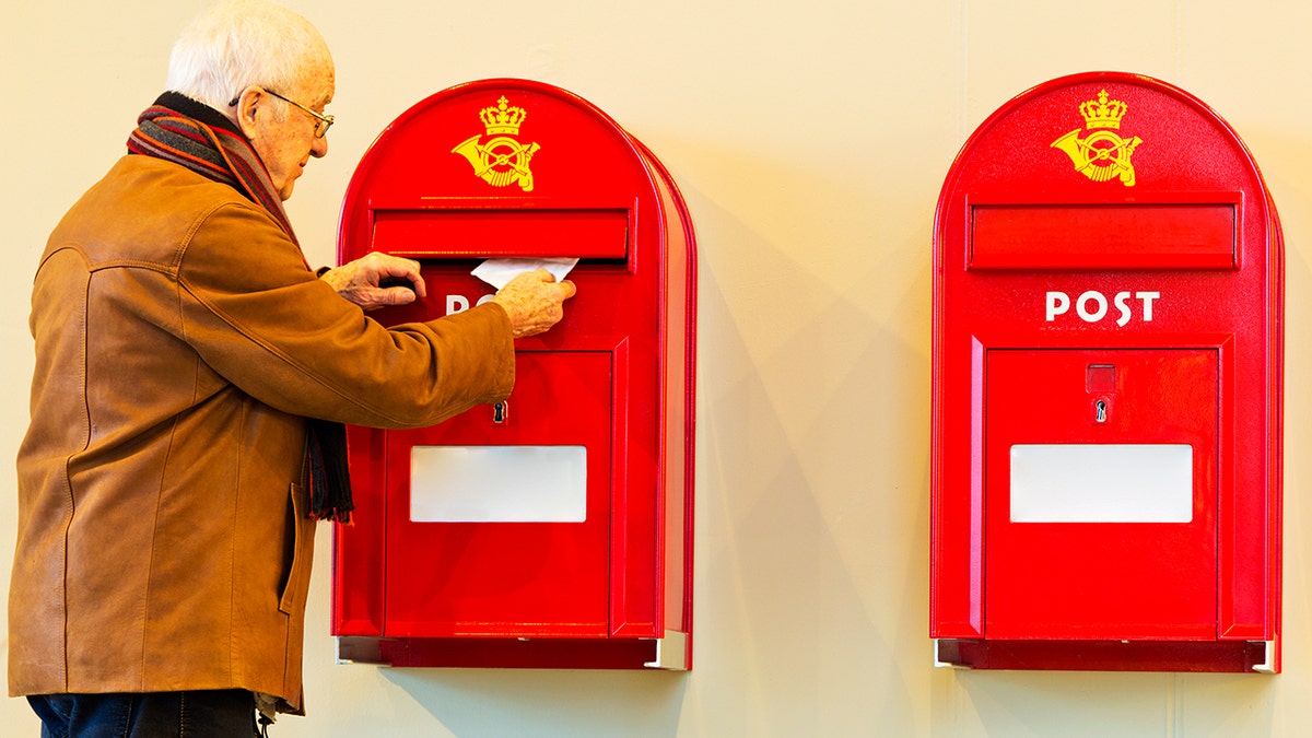 An elderly man wearing a brown jacket and a scarf around his neck puts a letter in one of two Danish mailboxes pictured next to each other.