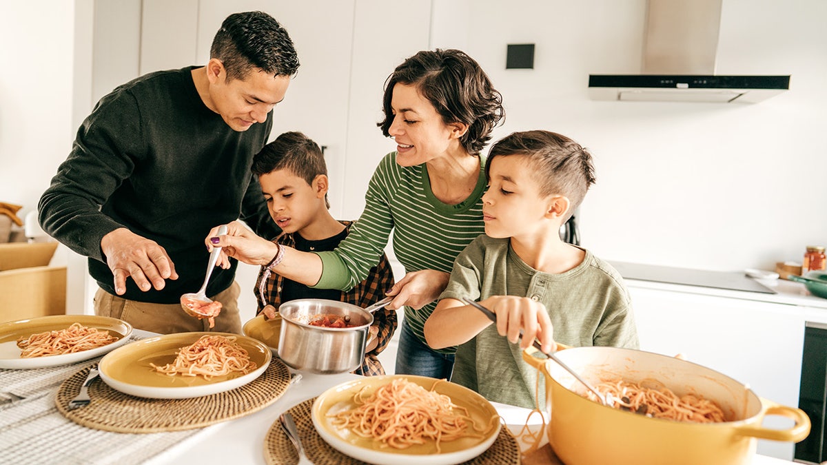 Mom, dad and two sons preparing freshly cooked pasta on plates at kitchen counter.