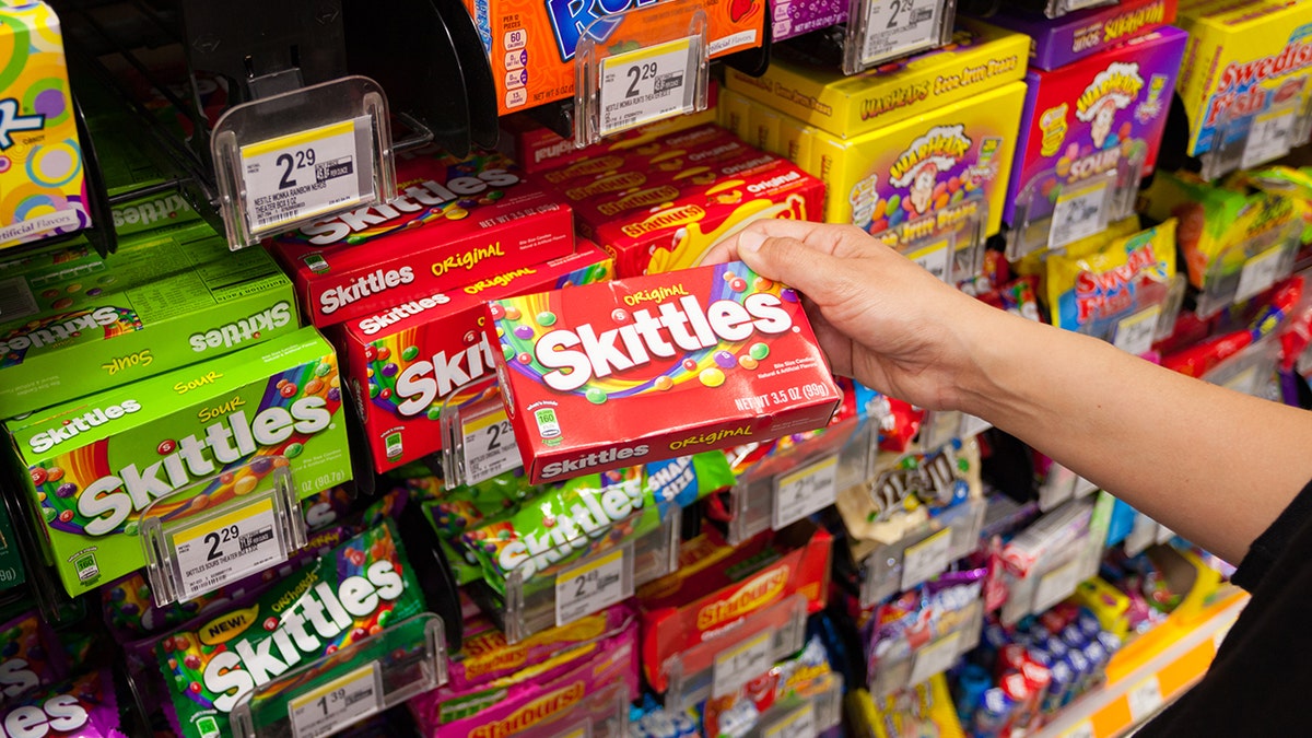 A mão de uma pessoa vista comprando skittles em uma vitrine de doces embalados e coloridos na loja.