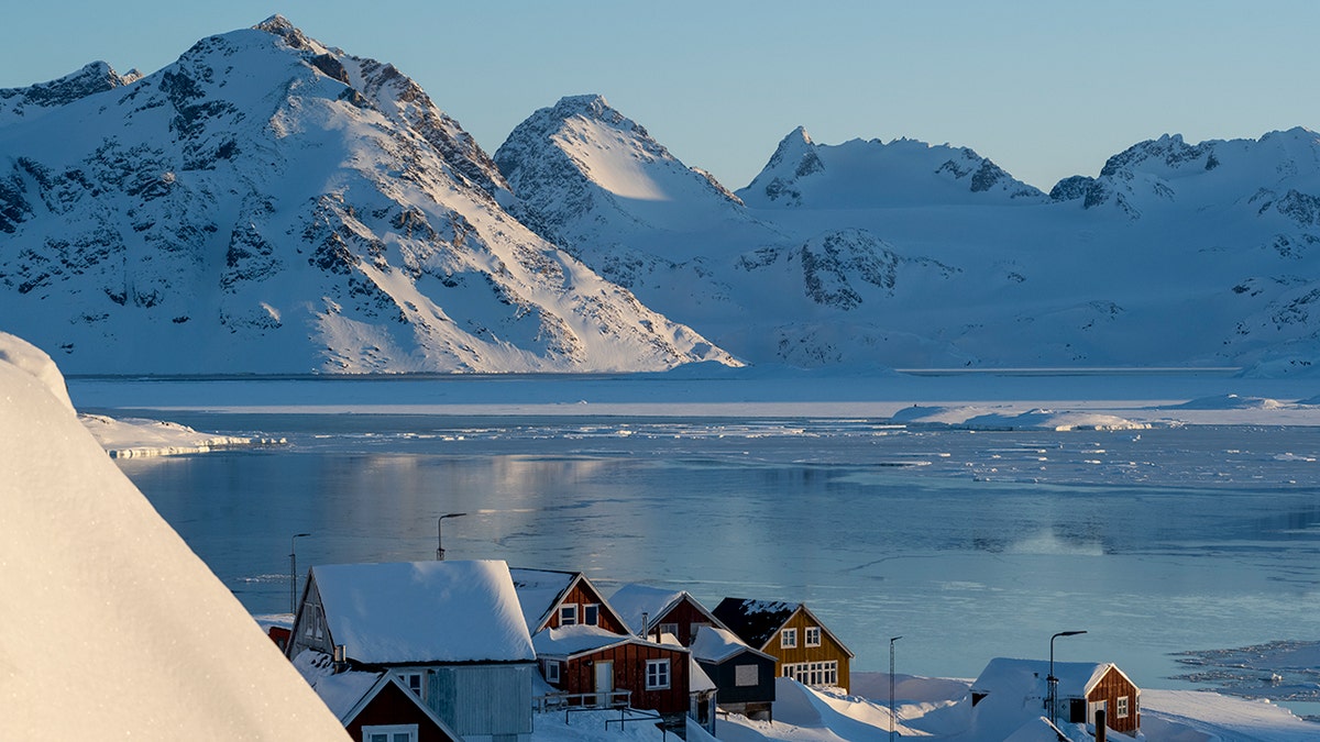 Snow-covered mountains rise above an icy Arctic fjord with small colorful houses along the frozen shoreline in Greenland.