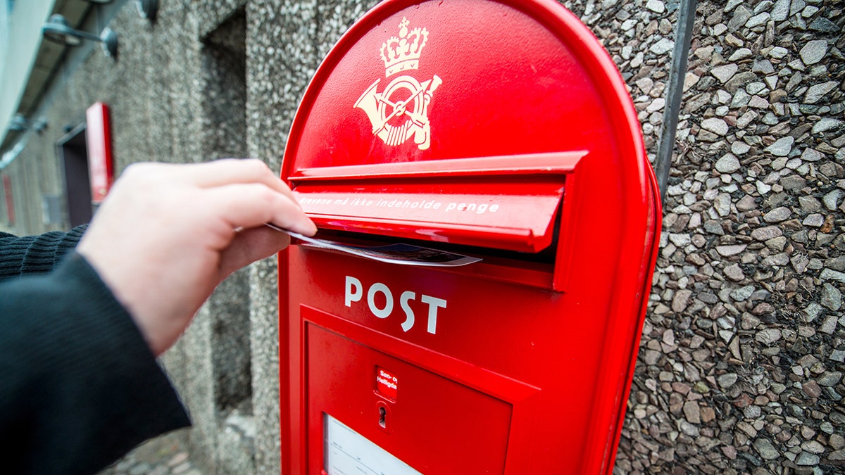 A hand drops a postcard into a red Danish mailbox.