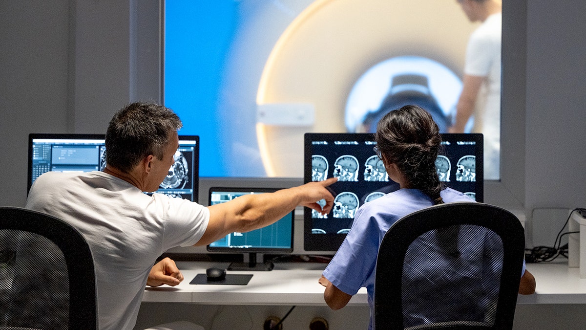 Rear view of male and female MRI technologist sitting at the console in the operating room and operating the MRI scanner