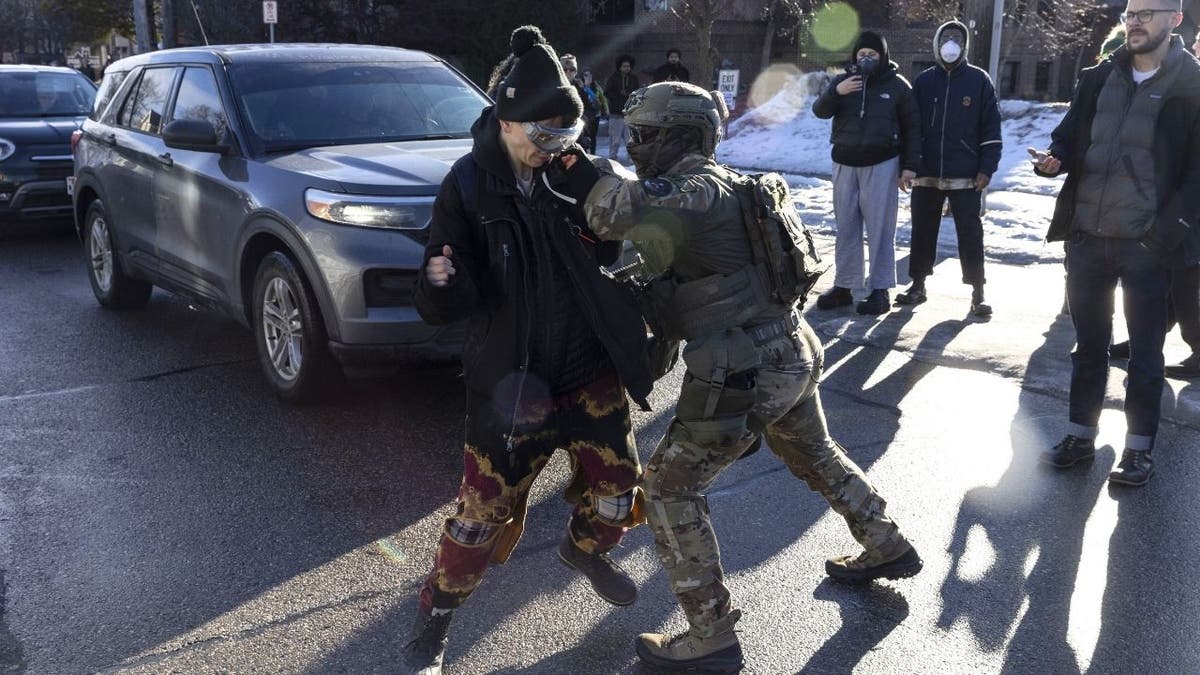 Federal agent in tactical gear pushes a person in the street with cars behind and bystanders watching.
