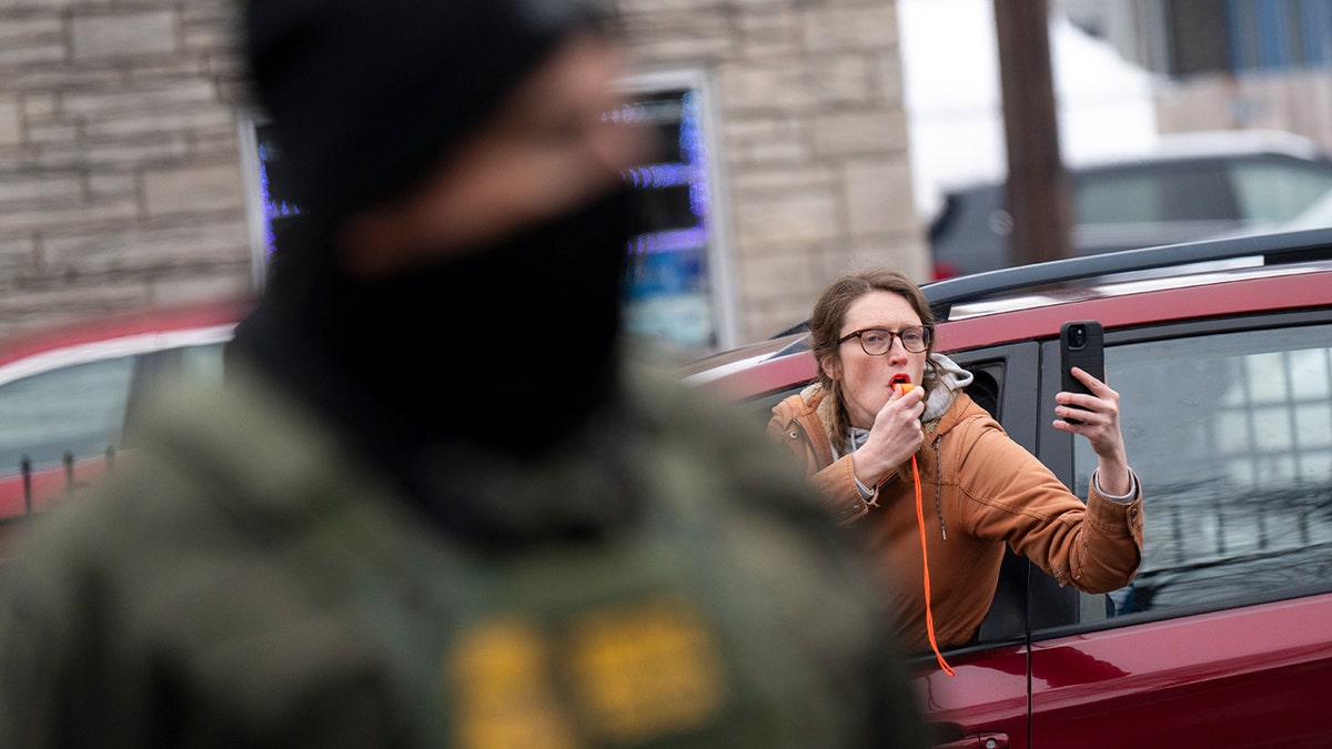 A bystander blows a whistle as federal agents detain a man on a city street.