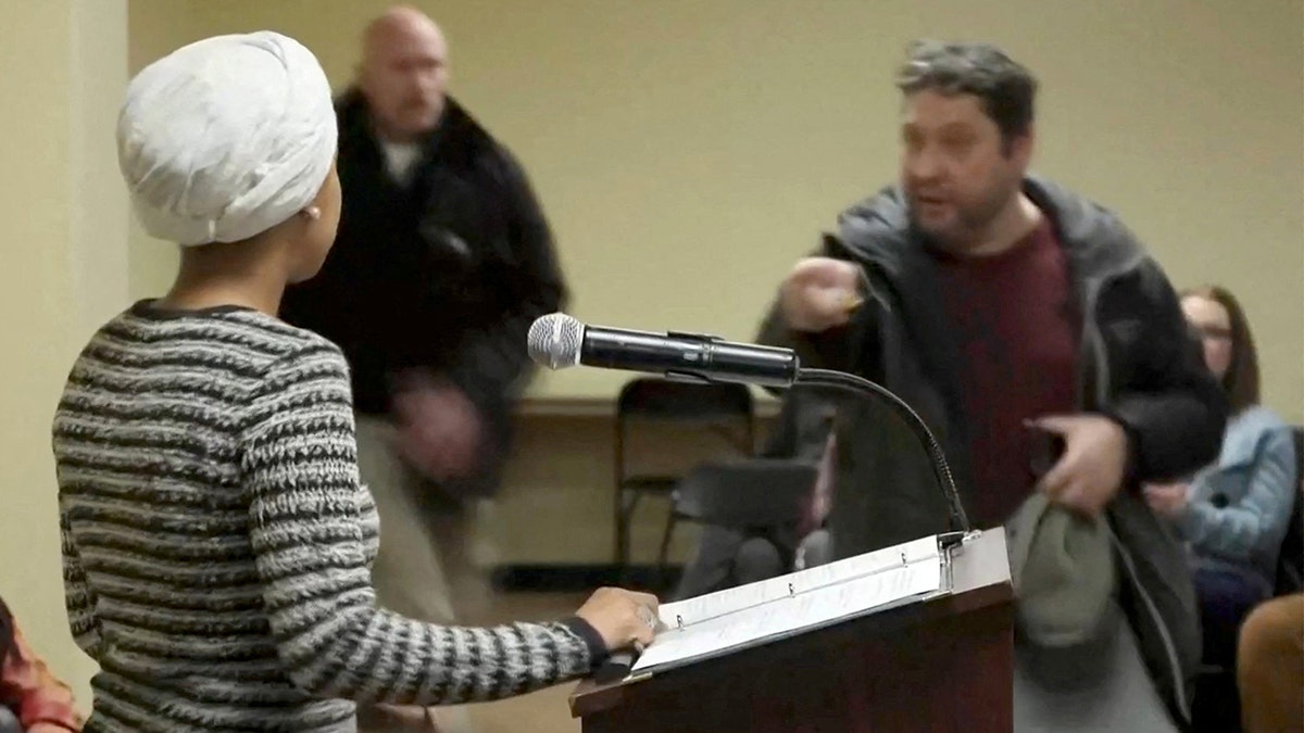 A man shouting and spraying a liquid toward Ilhan Omar during a town hall meeting.