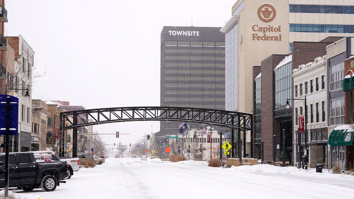 Snow falls along a downtown street lined with buildings and traffic lanes.