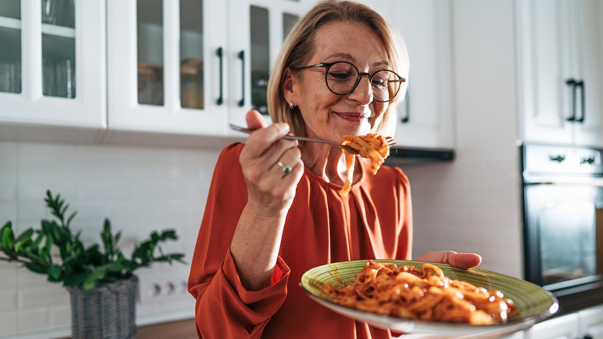 Older woman smiling as she eats pasta from plate twirled around fork in kitchen.