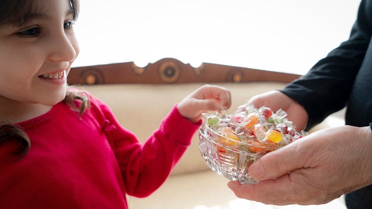 Little girl smiling as she takes piece of candy from older relative holding glass bowl of sweets for her.