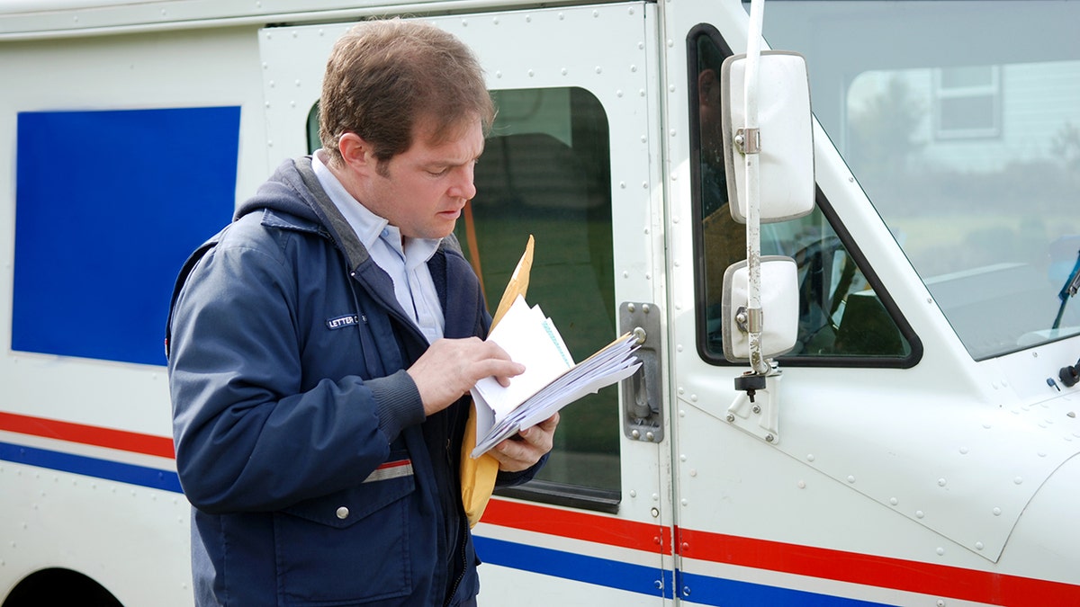 USPS mail carrier reviewing letters and packages beside a postal delivery truck.