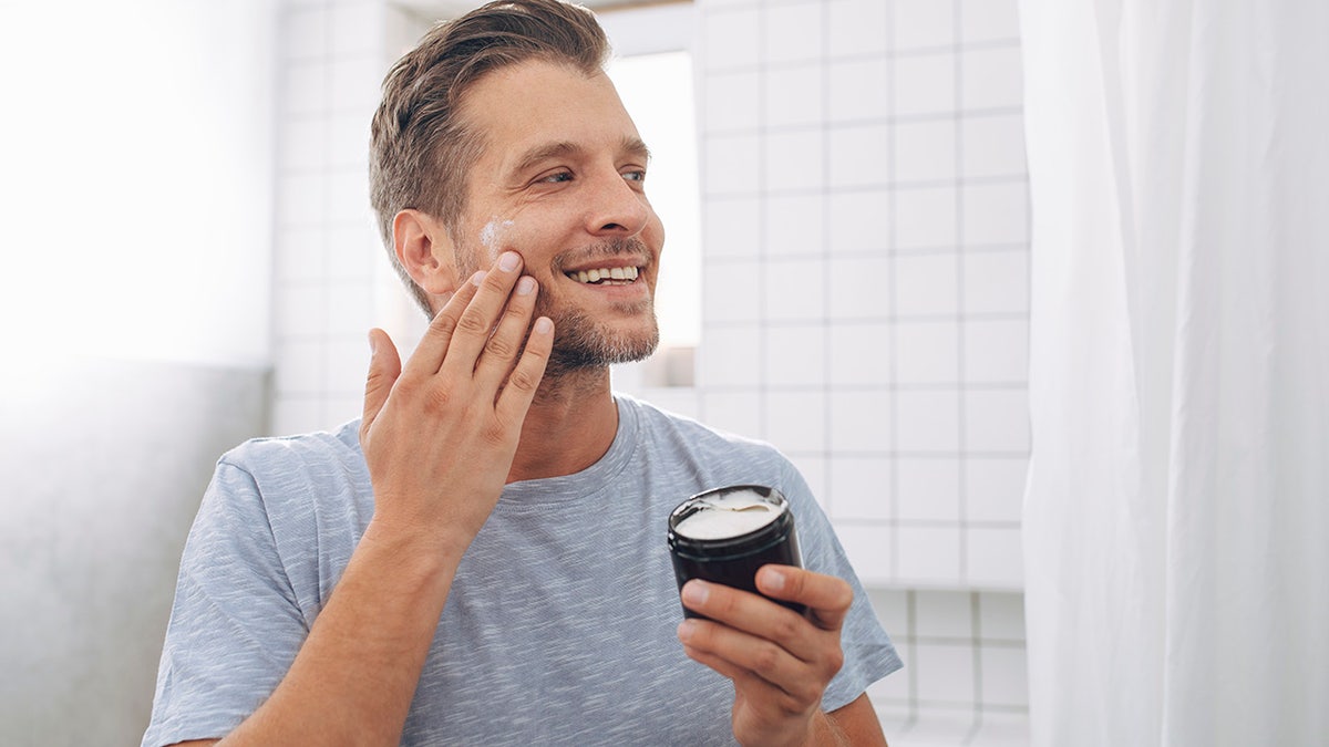 Young man applying lotion to face in mirror in bathroom, smiling as he holds jar of skincare product.