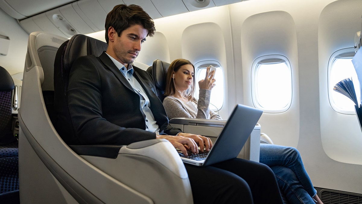 Airline passengers sitting in the cabin of an airplane, with a man working on a laptop and a woman using a smartphone during a flight.
