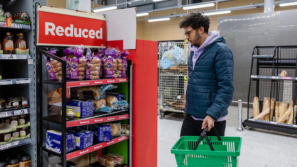Shopper holding a green basket while looking at reduced-price bread in a grocery store aisle.