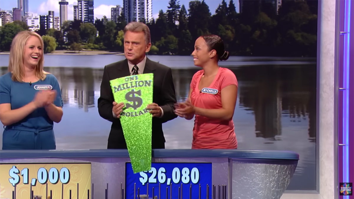 Pat Sajak holds a green prize card while two contestants applaud.