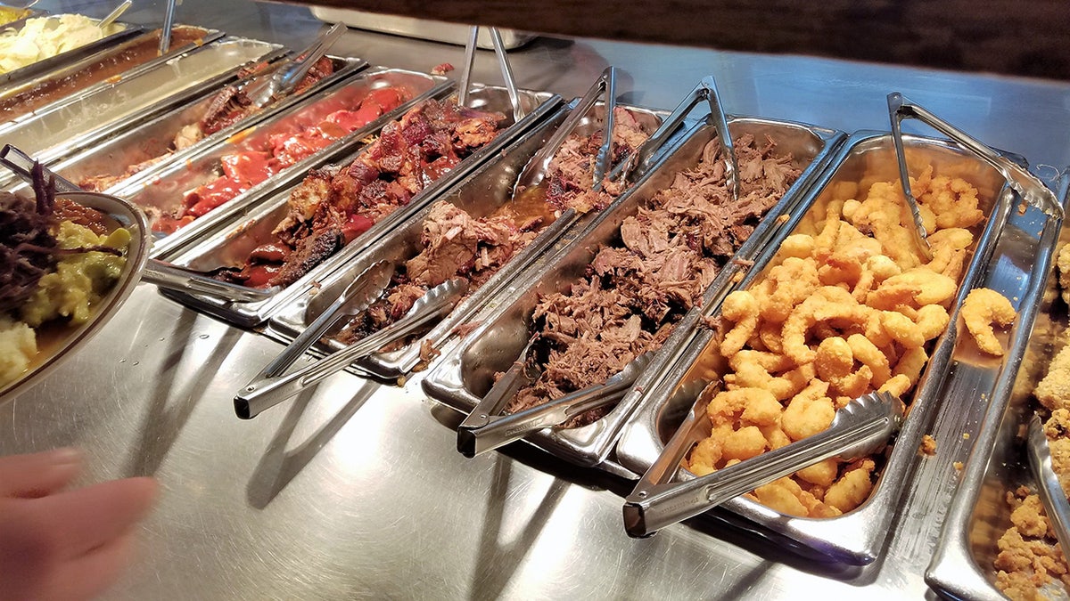 Buffet of hot food seen at Moonlite Bar-B-Q Inn in Owensboro, Kentucky as customer holding plate reaches for some.