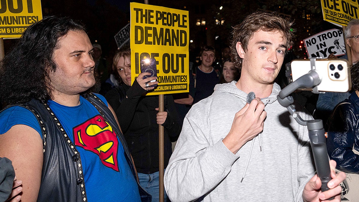 Nick Shirley holds a camera while filming protesters gathered during a demonstration on a city street.