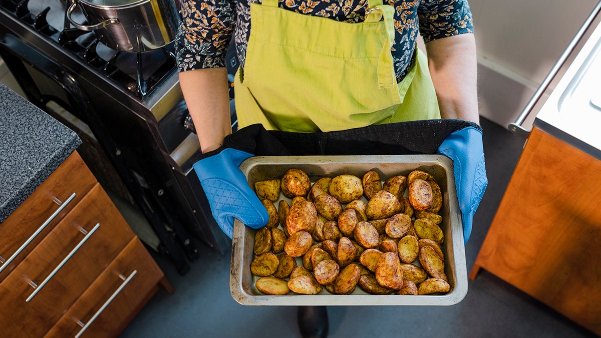 Woman holding plate of baked potatoes and wearing blue oven gloves in kitchen