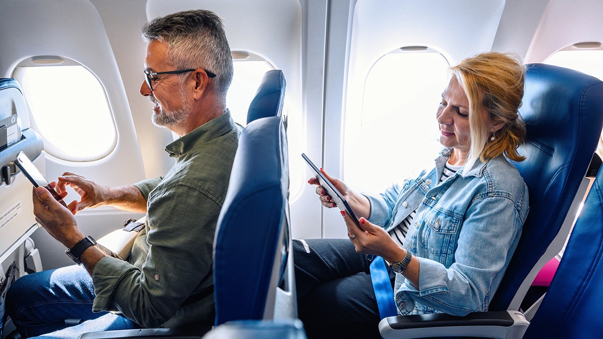 A man and woman sitting in their seats on the airplane, looking at their electronic devices.