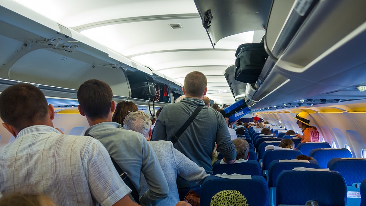 Passengers standing in a crowded airplane aisle during boarding with overhead bins filled with carry-on luggage.