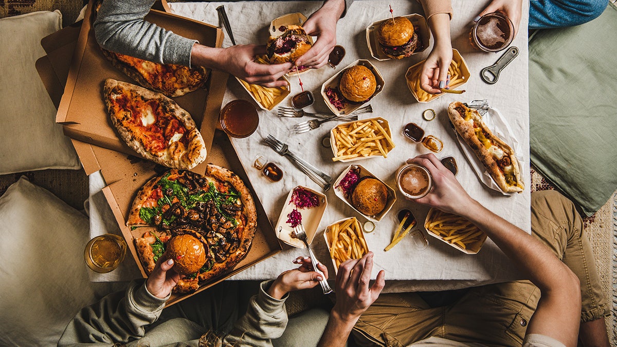 Friends eating different food delivery options in living room, including pizza, burgers and heroes.