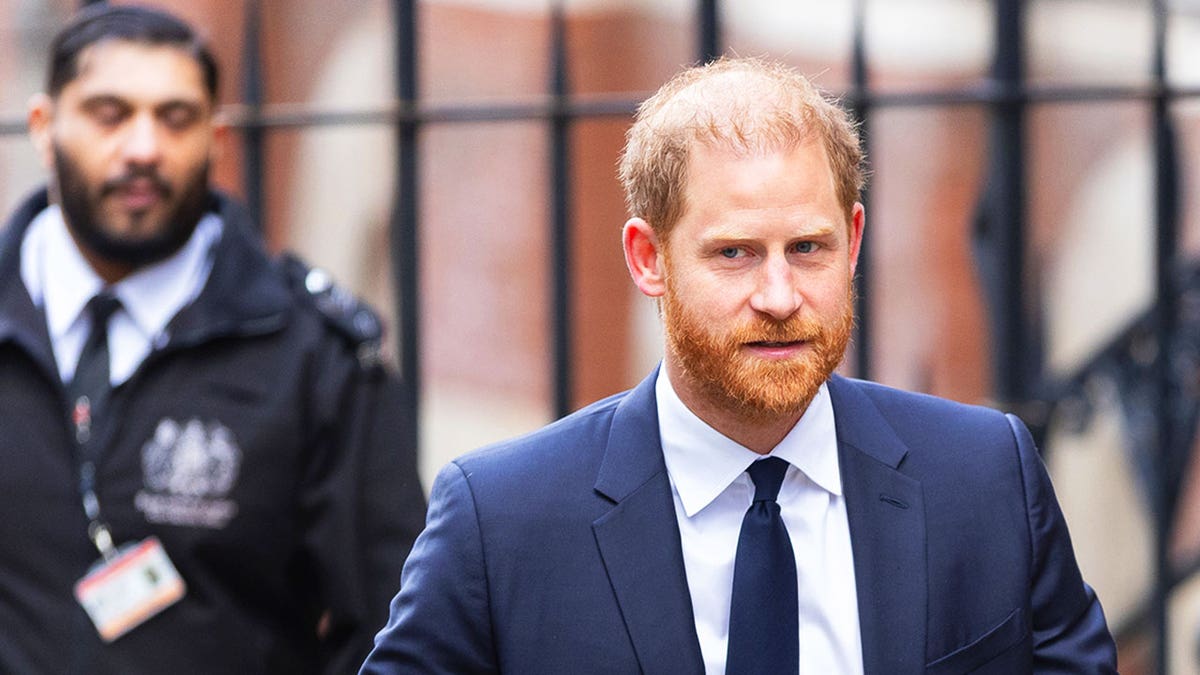 Prince Harry wearing a dark blue suit walking in front of a guard in London.
